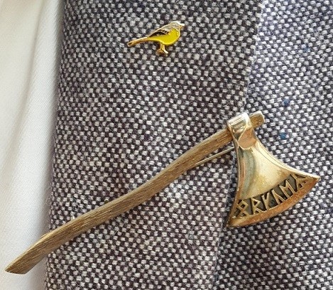 Gouden bijl-broche met runen-opschrift en een kleine gele vogel-pin op een grijs jasje — de strijdbijl van il partigiano.