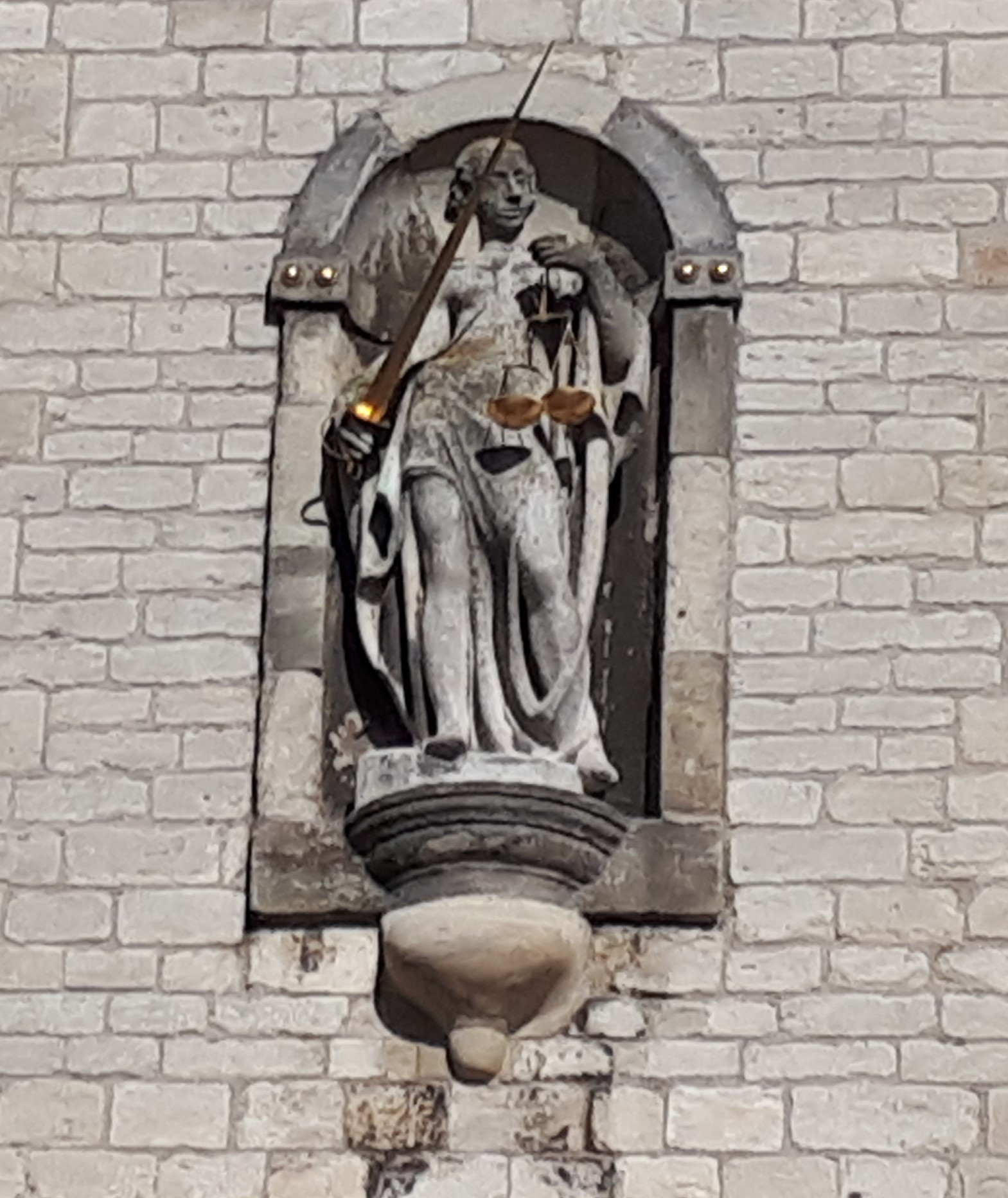 Stenen beeldje van Vrouwe Justitia met weegschaal en zwaard in een nis in een bakstenen gevel.